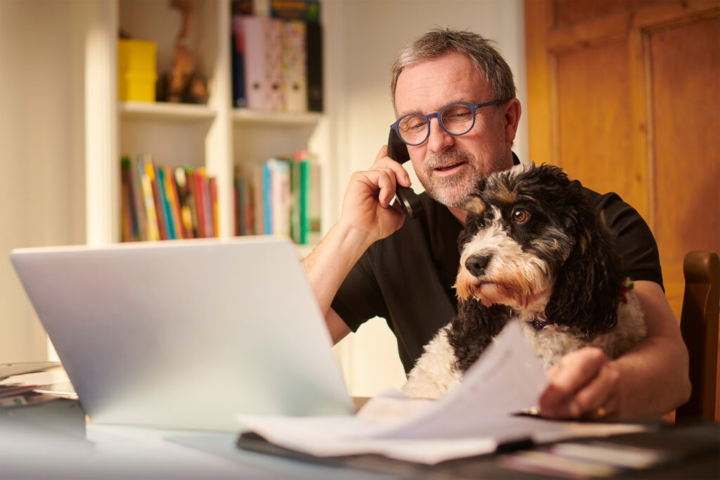 Man talking on the phone beside his dog at a computer deesk