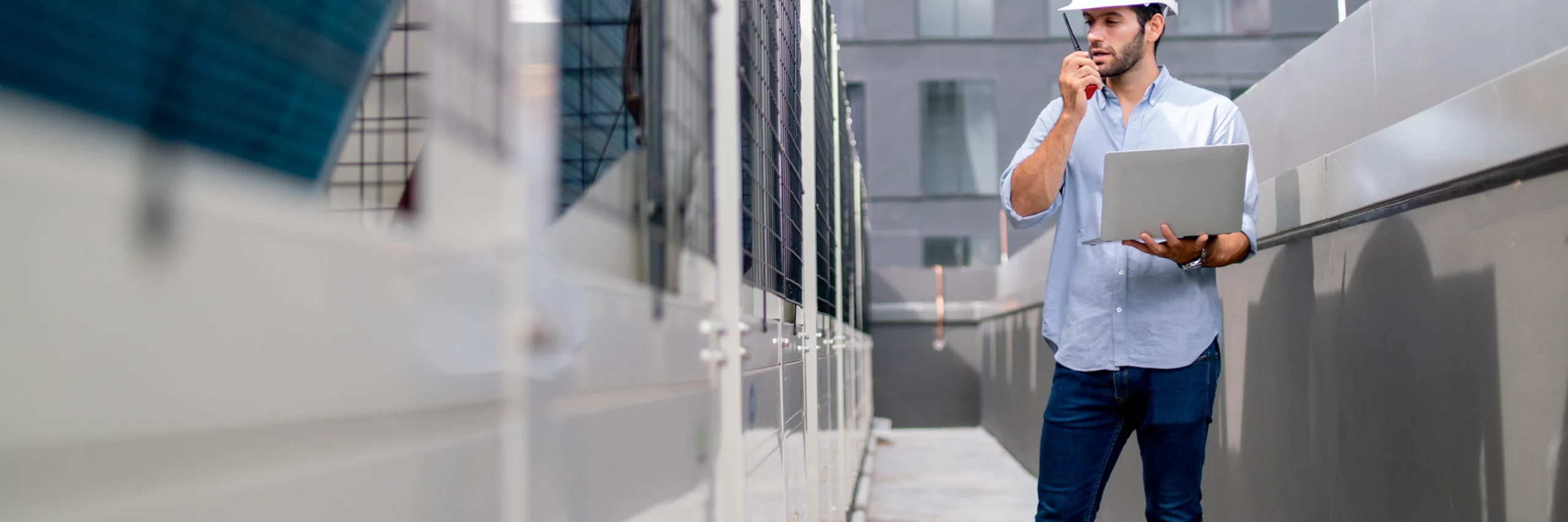 Man in white hardhat and blue button-up shirt carrying a laptop while making a call on a walkie-talkie